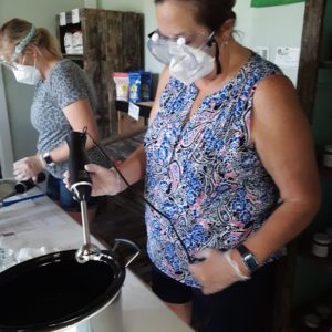 Two women wearing goggles and face masks use immersion blenders to mix soap ingredients in a crockpot. They attended the class as part of a mother and daughter event.