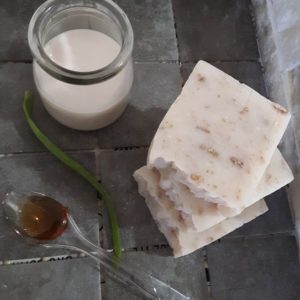 Three bars of natural soap sitting on gray stone. A spoonful of honey, a class of almond milk, a piece of aloe and a few grains of oatmeal are also on the gray stone.
