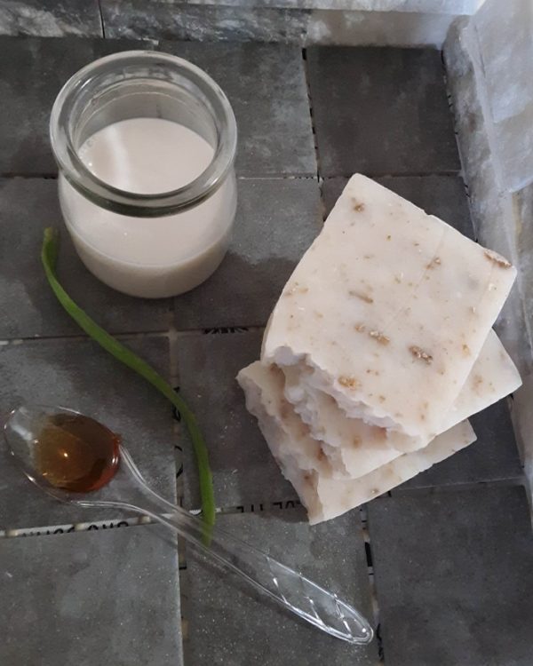 Three bars of natural soap sitting on gray stone. A spoonful of honey, a class of almond milk, a piece of aloe and a few grains of oatmeal are also on the gray stone.