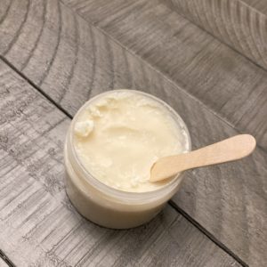 A clear jar of white deodorant cream with a wood application spoon sticking out of it.