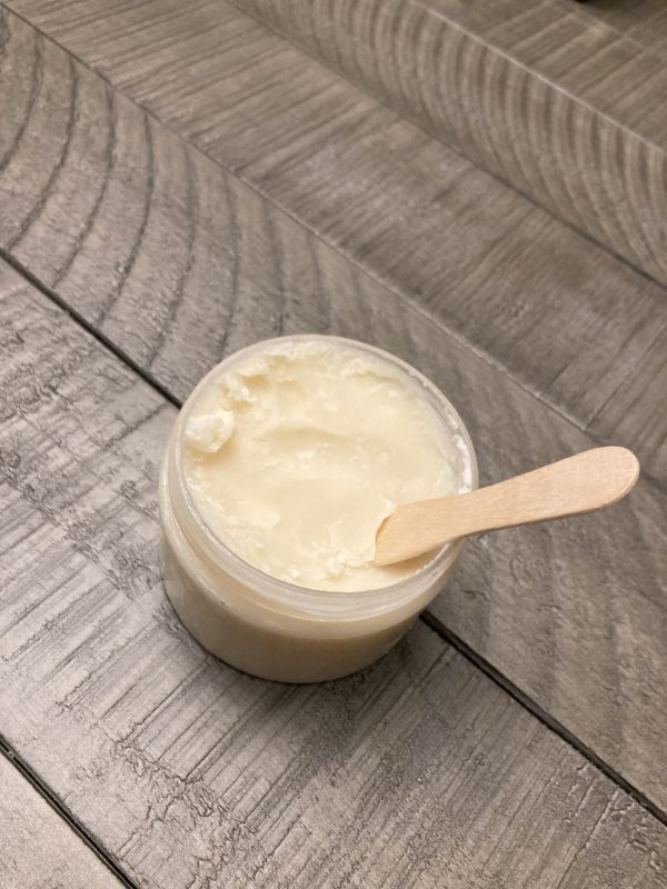 A clear jar of white deodorant cream with a wood application spoon sticking out of it.