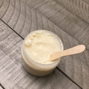 A clear jar of white deodorant cream with a wood application spoon sticking out of it.