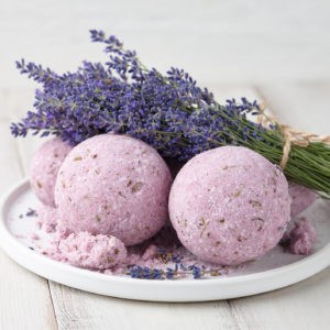 Three sphere-shaped bath bombs laying on a circular plate. The bombs are scented with lavender. A bunch of cut lavender is laying on top of them.