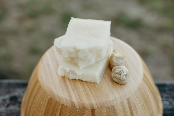 Three bars of natural off-white-colored soap sitting on an inverted wood bowl with a piece of ginger root beside them.