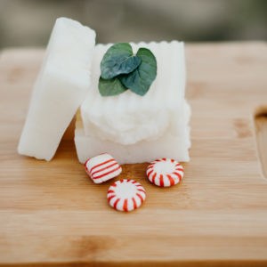 Three bars of natural soap with a mint leaf and peppermint candy beside them. All are sitting on a wood cutting board.