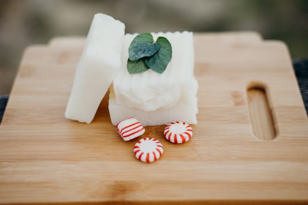 Three bars of natural soap with a mint leaf and peppermint candy beside them. All are sitting on a wood cutting board.
