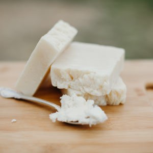 Three bars of off-white-colored natural soap with a spoon full of shea butter beside them. They are all sitting on a wood cutting board.
