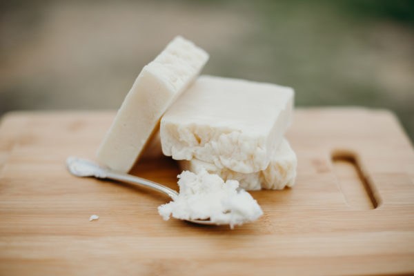 Three bars of off-white-colored natural soap with a spoon full of shea butter beside them. They are all sitting on a wood cutting board.