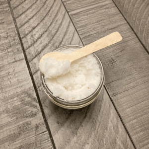 A clear glass jar of sugar scrub with a wooden spoon laying across the top of the open jar.