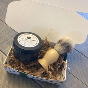 A decorative gift box containing a black circular can of shave soap. The box also contains a shave brush.