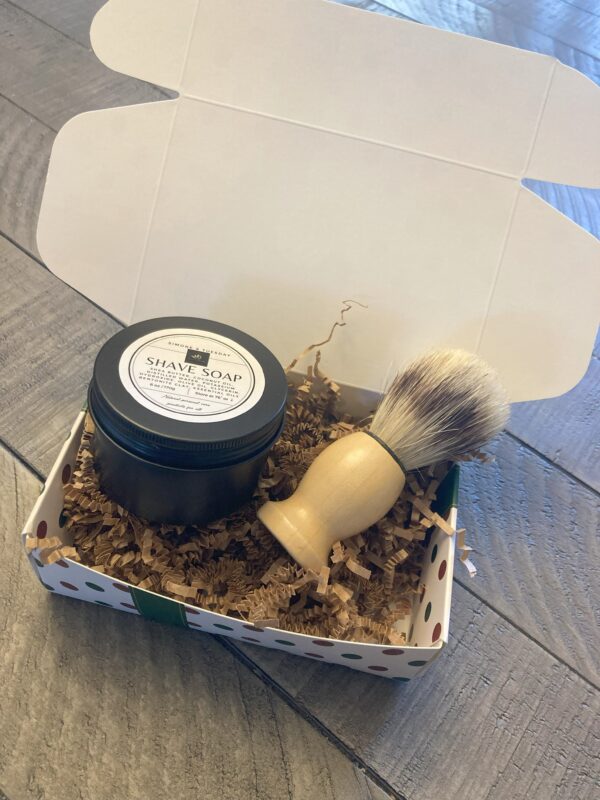 A decorative gift box containing a black circular can of shave soap. The box also contains a shave brush.